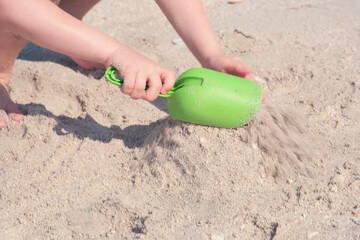 Hands of a child enthusiastically building sandcastle using a green toy shovel on a sandy beach....