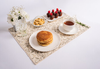 A cute and handmade breakfast on an individual flower tablecloth. There is a cup of tea and several dishes with pancakes and sweets, croissants, biscuits and a bouquet of white flowers
