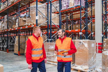 Two warehouse workers are discussing logistics while holding barcode scanners in a distribution center, surrounded by shelves full of goods