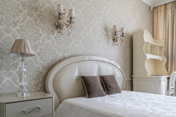 cozy retro-style bedroom with beige wallpaper, bed with pillows and bedspread, bedside table, lamp on table, wall lamp. bedroom interior. vintage chandelier with crystals, chest of drawers.