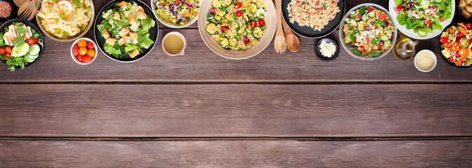 Variety of delicious homemade salads. Above view top border on a dark wood banner background. Summer food concept. Potato, caesar, pasta and green salads.