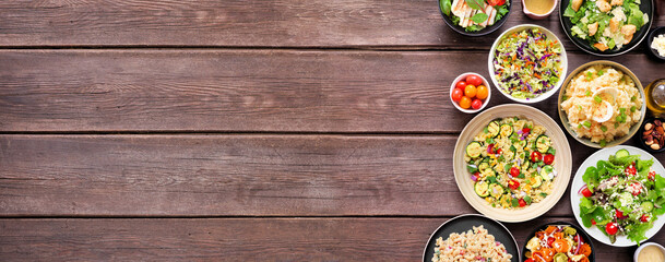 Variety of delicious homemade salads. Above view side border on a dark wood banner background. Summer food concept. Potato, caesar, pasta and green salads.