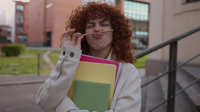 Cheerful redhead student with curly hair playfully wearing pencil as mustache, holding notebooks, smiling brightly while expressing lighthearted academic joy on campus
