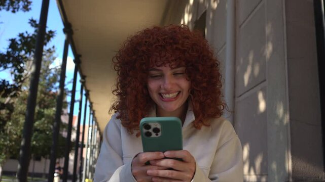 Young woman with curly red hair enjoying sunny campus day, laughing while texting on smartphone under architectural portico with casual, genuine energy