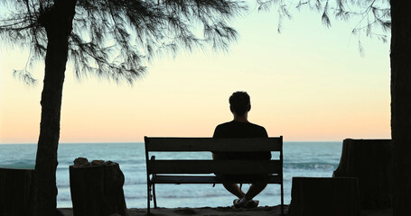 Silhouette of man sitting on bench by sea under trees at sunset.
