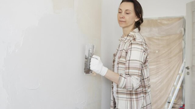 Woman applying plaster to wall with putty knife, wearing protective gloves and creating smooth surface during home renovation work