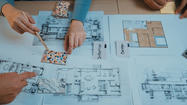 Hands of designers actively engaging in a collaborative session to select color swatches and analyze architectural plans on the table for an interior design project. SACTR