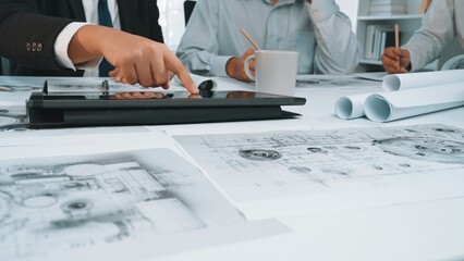 A diverse business team engages in an architectural project, utilizing a digital tablet while reviewing detailed blueprints and designs in a modern office setting. SACTR