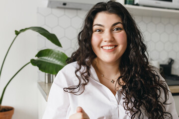 Indoor portrait of happy surprised video blogger or vlogger sharing exciting story of her life or tips of losing weight and her inspiring results, sitting at kitchen table and smiling at camera