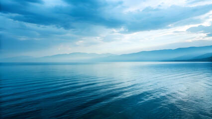 Obraz premium Serene Blue Landscape Water Ripples Hazy Mountains and Dramatic Sky.