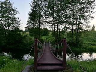 Rustic wooden footbridge suspended over a tranquil stream, surrounded by lush green trees and grass. The wooden planks are dark, secured with sturdy cables. Peaceful, pastoral setting