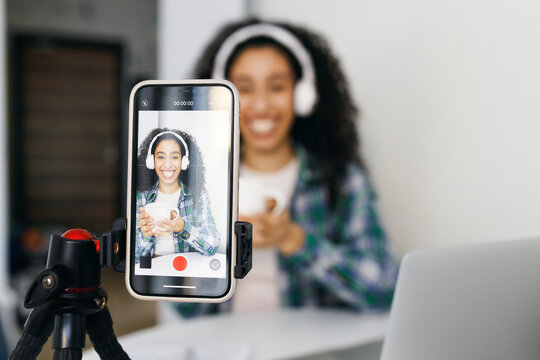 Selective focus on smartphone mounted on tripod with screen displaying happy cheerful generation z female of african american ethnicity in headphones recording video blog drinking coffee or tea