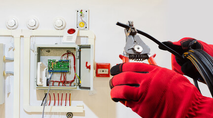 Electrician in protective gloves installs or repairs a fire alarm control system, preparing wires for connection, related to electrical and safety systems work.