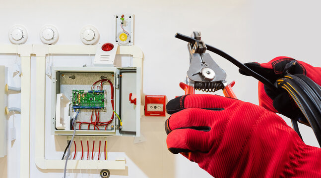 Electrician in protective gloves installs or repairs a fire alarm control system, preparing wires for connection, related to electrical and safety systems work.