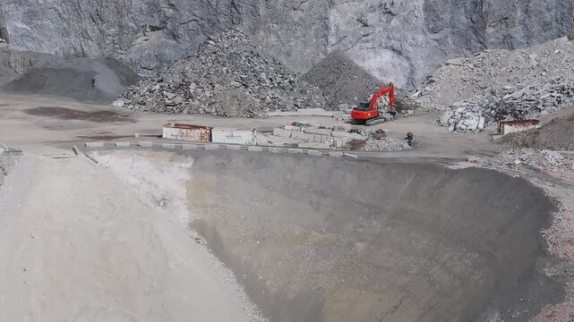 Aerial view of a mining site in the Alps showing a red excavator in motion, rugged terrain, steep cliffs, and a winding dirt road through the quarry.