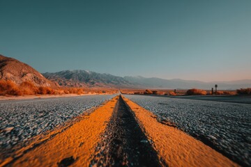 Long straight road stretching towards distant snow-capped mountains under clear blue sky, orange roadside bushes lining the asphalt, leading the eye to a serene landscape.