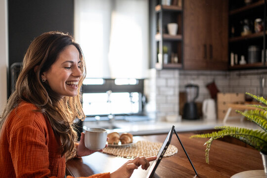Happy woman using tablet and drinking coffee in modern kitchen