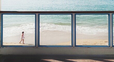 A person walking on the sand of the beach behind the glass walls of the promenade with sunlight and the sea in the background.