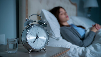 Alarm clock on bedside table, woman sleeping in background, glass of water nearby, suggesting bedtime routine or sleep concept