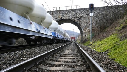 Low-angle view of a moving freight train on railway tracks passing under a stone arch bridge,...
