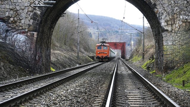 Red freight train passing under a stone railway bridge on double tracks in a mountainous rural area, emphasizing transportation and infrastructure.

