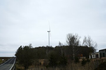 A tall wind turbine stands against a cloudy sky beside a gently curving road through farmland.