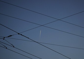Crisscrossing power lines in the sky with a plane's contrail cutting across, highlighting structure and motion.