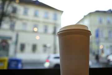 A white paper coffee cup stands in focus with a soft blurred view of buildings behind.