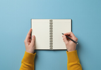 Obraz premium Top-down flat lay view of hands wearing a yellow sweater, writing with a black pencil in a spiral notebook on a minimal light blue desk, symbolizing work, study, or planning