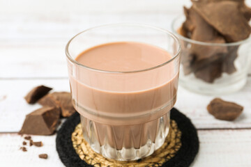 Tasty chocolate milk in glass and pieces of chocolate on white wooden table, closeup