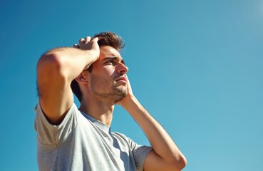 Naklejka premium Young man suffers heat stroke outdoors. Guy holds head hands against background clear blue sky. Overheating causes cephalalgia, health risks during summer vacation, sunny hot weather, dehydration.