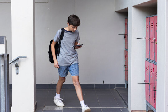 Walking in school hallway, boy with backpack using smartphone near lockers