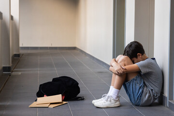 Sitting in school hallway, boy hugging knees with backpack and notebooks nearby