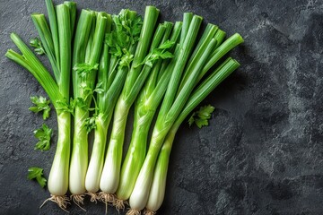 Obraz premium Fresh green onions arranged neatly on a dark stone surface with herbs in the background