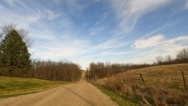 Car point of view traveling on a single lane rural gravel road in the Midwest, USA . The road winds up and down hill. Views of empty agricultural fields and farm houses. Big blue cloudy skies above.