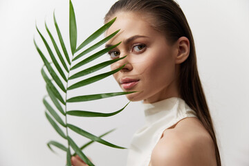 Naklejka premium Elegant portrait of a young woman holding a palm leaf, showcasing natural beauty and serenity in a minimalist setting with soft colors.