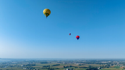 Brightly colored hot air balloons ascend into a clear blue sky, showcasing an expansive view of lush green fields and small villages below on a beautiful autumn day.
