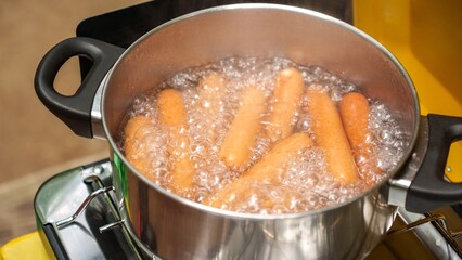 A stainless steel pot filled with boiling water and cooking sausages on a camping gas burner.