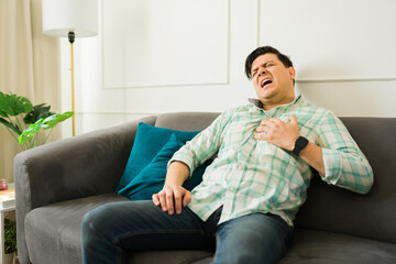 Hispanic man having a heart attack and touching his chest while experiencing painful angina and shortness of breath in his living room