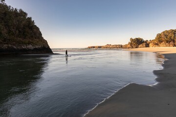 A person paddleboards in calm waters at a beach in Mendocino, California, USA. The clear blue sky and tranquil scene evoke a sense of peace and outdoor adventure.