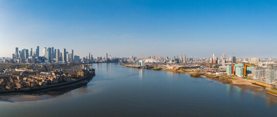 Naklejka premium Aerial view of London with the River Thames, Canary Wharf skyscrapers, O2 Arena, residential areas, green spaces, and a clear blue sky.