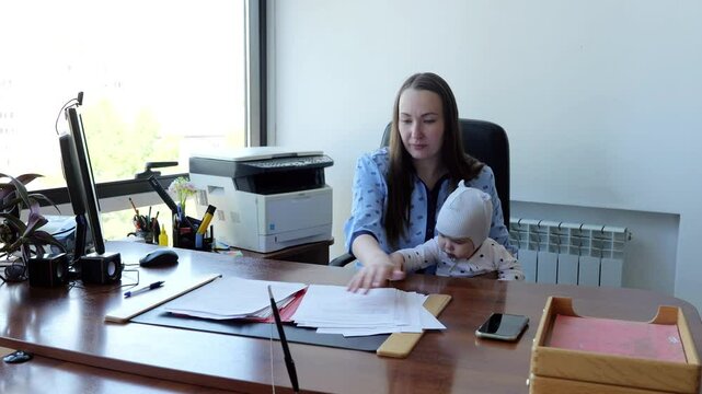 Businesswoman seated in office managing tasks while holding her baby juggling work and childcare. Successful caucasian woman works in office multitasking with infant in her arms working mother.