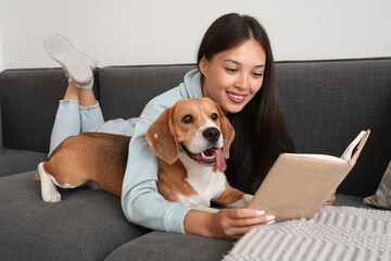 Young woman reading book and lying with adorable Beagle dog on sofa in living room