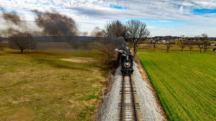 A vintage steam locomotive chugs along the railway track, emitting smoke as it travels through a serene rural landscape with green fields and bare trees under a blue sky.