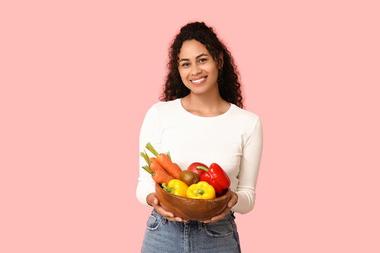 Young happy African-American woman with bowl of different fruits and vegetables on pink background. Independence from Meat Day