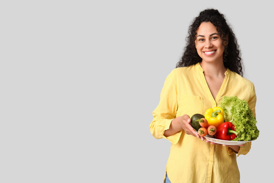 Young happy African-American woman with plate of different vegetables on grey background. Independence from Meat Day
