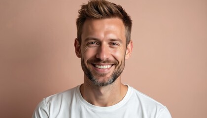 Fototapeta premium Portrait of attractive caucasian man in white t-shirt smiling. Happy male with stylish hair looking at camera on neutral beige-pink background. Forty years old. Studio shot. Copy space.