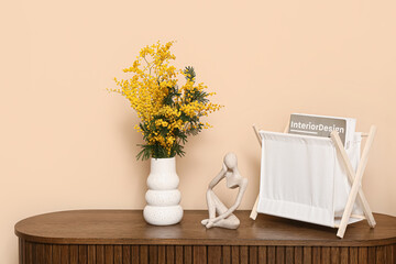 Vase with mimosa flowers, statuette and magazine stand on shelf near beige wall