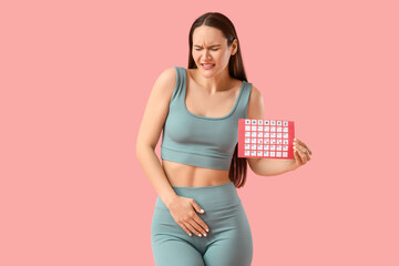 Sporty young woman with menstrual calendar suffering from cramps on pink background