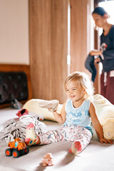 Fototapeta premium Little girl with a sneaker in her hand sits on a bed among toys